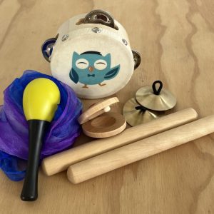 An assortment of musical instruments including a wooden owl tambourine, a yellow maraca on a purple scarf, wooden castanets, brass finger cymbals, and rhythm sticks