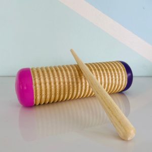 A cylindrical wooden guiro with pink and purple painted ends, placed next to a wooden scraper stick