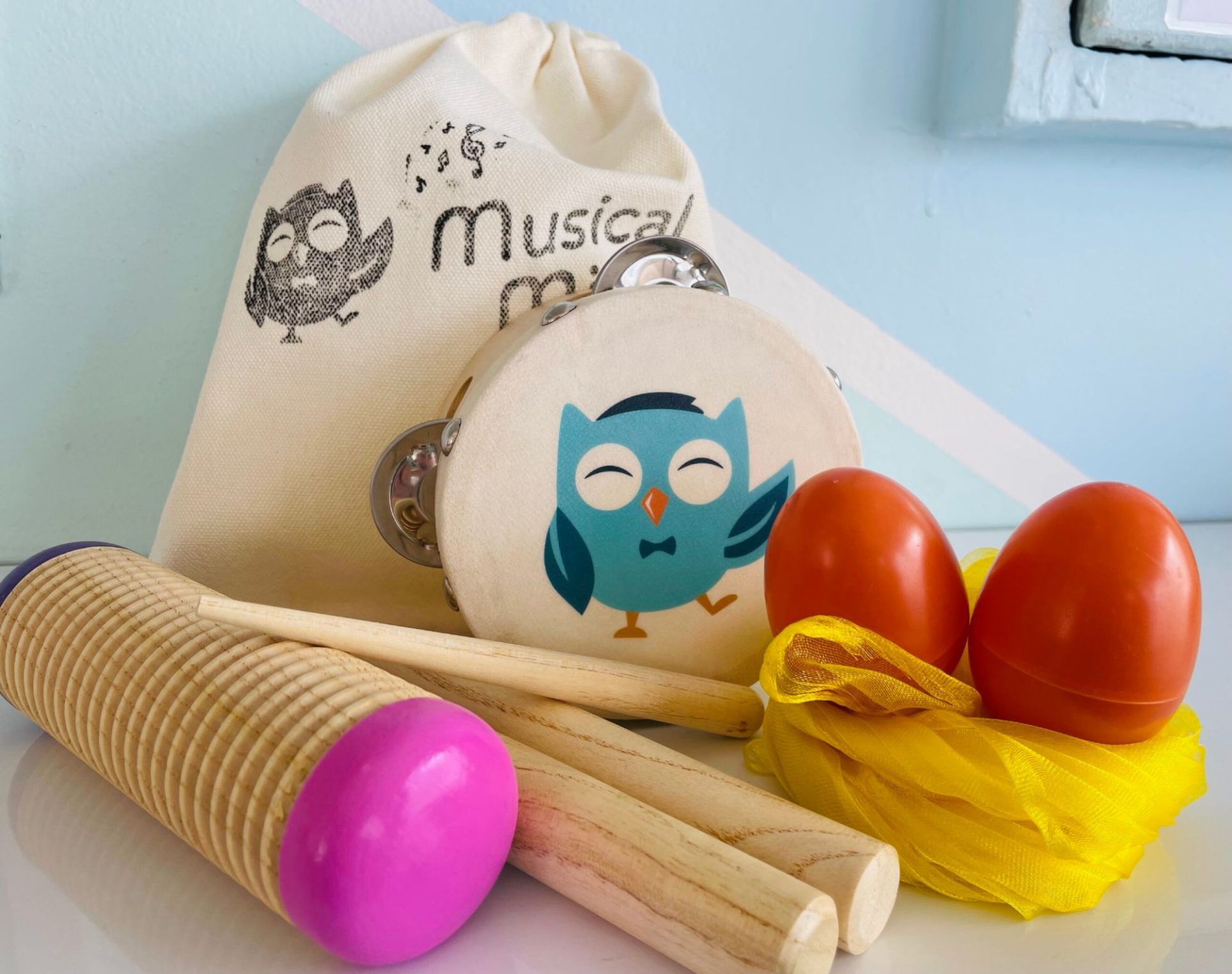 A rhythm instrument pack featuring a canvas bag, a wooden tambourine with a blue owl, two orange egg shakers on a yellow scarf, a wooden guiro, and rhythm sticks