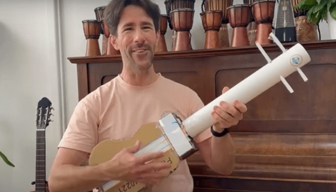 A smiling man holding a handmade string instrument constructed from cardboard and a PVC pipe, standing in front of a piano and several djembe drums